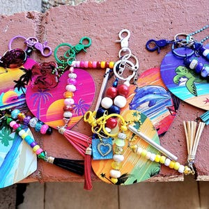May include: A collection of heart-shaped keychains with tropical sunset and beach designs. Each keychain features colorful beads, tassels, and metal clasps. The keychains are adorned with palm trees, turtles, and other beach-themed elements.