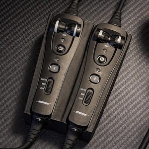 May include: Two black Bose audio control modules with attached cables. Each module has a volume control, mute switch, and power button. The modules are connected and sit on a textured surface. The Bose logo is visible.