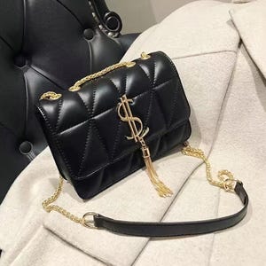 May include: Black quilted crossbody bag with a gold chain strap and a gold dollar sign emblem. The bag features a flap closure and a gold tassel. The bag is made of a smooth, shiny material.