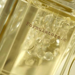 May include: A clear, rectangular perfume bottle with a light yellow liquid inside. The text "Formulators.c" is printed on the front. The bottle has a unique design with bubble-like shapes. The background is a soft, blurred white.