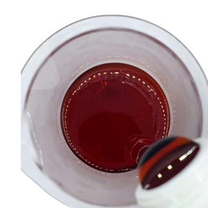 May include: A clear glass beaker filled with a deep red liquid, possibly a solution or a beverage. A white bottle with a black and red striped top is partially visible, suggesting pouring. The image is taken from a top-down perspective.