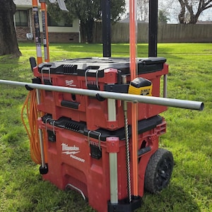 May include: Red Milwaukee Packout rolling tool storage system with various tools attached. Includes a level, conduit benders, and other equipment. The system is on a grassy lawn.