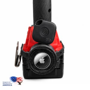 May include: A black Tile tracker and an Apple AirTag are shown on a red and black object. The Tile tracker has an @ symbol and the word "tile". The AirTag has the Apple logo. The image also includes the text "Designed and Manufactured in AMERICA" with an American flag.