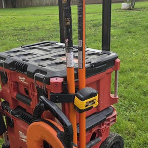 May include: A red and black rolling tool chest with various tools attached. Includes orange conduit benders, a yellow and black tape measure, and an orange fish tape. The tools are on a green grassy lawn.