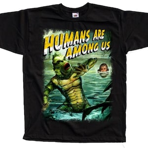 May include: A black short-sleeve t-shirt featuring a graphic print of a green, scaly creature emerging from water toward a person in the distance. The text HUMANS ARE AMONG US is printed in large yellow and white letters above the creature.