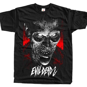 May include: Black t-shirt featuring a detailed, grayscale image of a monster face with glowing eyes and red accents. The text "EVIL DEAD 2" is printed in white below the image. The shirt is made of a soft material.