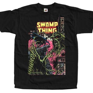 Może przedstawiać: Czarna koszulka z grafiką Swamp Thing i postacią przypominającą nietoperza, z napisem "SWAMP THING" w kolorze żółtym i czerwonym. Projekt zawiera krajobraz miejski z różowymi i zielonymi akcentami, tworząc styl komiksowy.