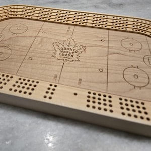 Pode incluir: Um tabuleiro de cribbage de madeira com um design de pista de hóquei. O tabuleiro apresenta um logotipo de folha de bordo com o texto "TORONTO MAPLE LEAFS". O tabuleiro tem várias filas de buracos para pregar e uma borda curva.