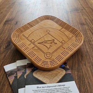 Może przedstawiać: Ręcznie wykonana plansza do gry w tryktraka z klonu z motywem diamentu baseballowego i logo Blue Jays. Plansza jest grawerowana z układem boiska do baseballu i ma liczne otwory na kołki. Deska ma około 30 cm długości.