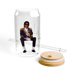 Puede incluir: Un vaso transparente en forma de lata con una imagen de Michael Jackson. La imagen muestra a Jackson con un traje negro, gafas de sol y una chaqueta dorada. Se incluye una tapa de madera y una pajita de cristal.