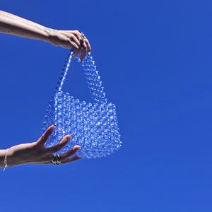 May include: A small, rectangular handbag constructed entirely from translucent, bright blue spherical beads. The bag features a structured body and two matching beaded handles, with the light reflecting off the glossy surfaces of the beads.