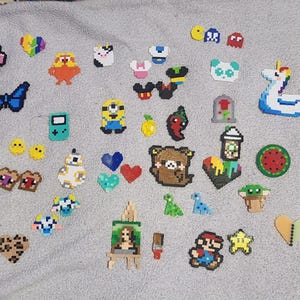 Puede incluir: Una colección de creaciones de arte pixelado coloridas, que incluyen un personaje rosa de Among Us, una mariposa azul, una carita sonriente amarilla, una consola de juegos verde, un Minion, un oso, un unicornio y una figura de Mario. Son decoraciones.