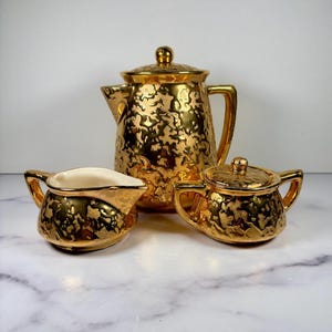 May include: A three-piece tea set featuring a coffee pot, a creamer, and a sugar bowl with a lid. Each piece is finished in a reflective metallic gold glaze with a textured, mottled pattern. The interior of the creamer is a smooth, off-white color.