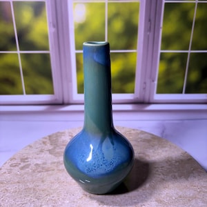 May include: A handcrafted ceramic vase with a rounded base and a long, slender neck. The vase features a gradient of colors, transitioning from a deep teal at the base to a vibrant blue at the top. The vase is displayed on a stone surface.