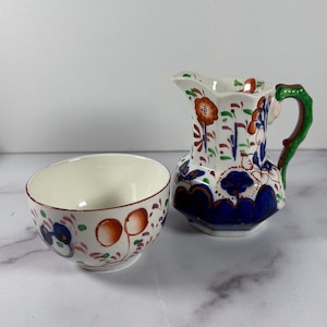 May include: A white ceramic creamer pitcher and bowl set. The pitcher has a green handle and is decorated with orange and blue floral patterns. The bowl has a similar design with orange and blue floral patterns. The set is on a white surface.