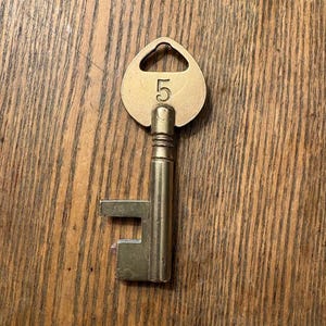 May include: A vintage brass key with a rounded top and the number "5" engraved on it. The key has a rectangular bit and a cylindrical shaft. The key is set against a wooden background.