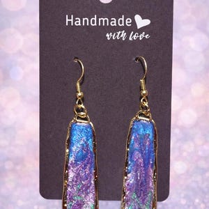 May include: Handmade earrings with a gold-tone frame and a rectangular, iridescent center in shades of blue, purple, and green. The earrings hang from gold-colored hooks. The black tag behind the earrings reads "Handmade with love."