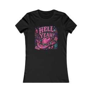 May include: A black short-sleeve t-shirt featuring a graphic print of a pink cowboy hat centered below the text HELL YEAH!. The design includes colorful string lights, a guitar, a cactus, a vinyl record, a star, and a small sign reading LIVE LOUD Y'ALL.