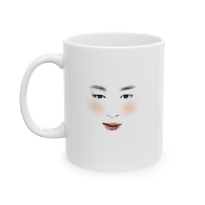 May include: A white ceramic mug featuring a minimalist graphic illustration of a face. The design includes dark eyebrows, detailed eyes, soft pink blush on the cheeks, and reddish-brown lips centered on the front of the smooth, glossy white surface.