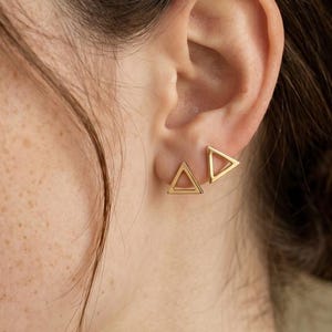 May include: A pair of gold-toned stud earrings, each featuring a small, open-frame triangle design. The earrings are worn on the earlobe, showcasing a polished metallic finish and a geometric shape that sits flush against the skin.
