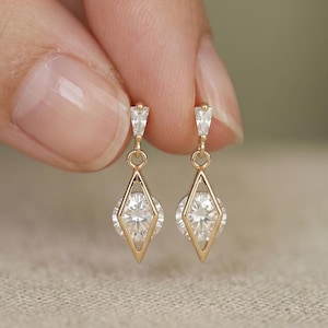 Può includere: Un paio di orecchini pendenti in oro con una pietra rotonda taglio brillante incastonata in una cornice geometrica a forma di diamante. Ogni orecchino è sormontato da una piccola pietra triangolare fissata a un sottile perno in oro.