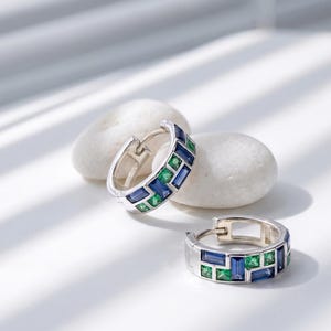 May include: A pair of silver hoop earrings featuring a geometric pattern of baguette-cut blue sapphires and square-cut green emeralds set into the metal band. The earrings have a hinged closure mechanism and a polished metallic finish.
