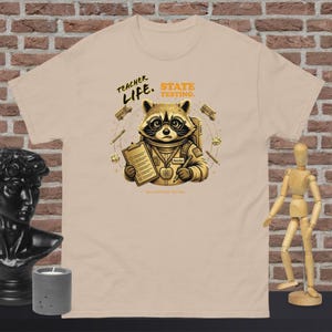 Peut inclure: T-shirt beige avec un raton laveur de dessin animé portant des lunettes et un uniforme d'enseignant, tenant un presse-papiers. Le t-shirt porte le texte "Teacher Life. State Testing." et des graphiques de bus scolaires. Le t-shirt est en matière souple.