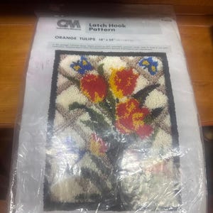 May include: A Columbia-Minerva latch hook pattern kit titled Orange Tulips, measuring 18 inches by 24 inches. The package contains a printed canvas featuring a floral design with red and yellow tulips, green leaves, and blue butterflies on a white grid.