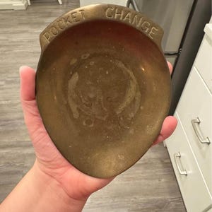 May include: A tarnished brass pocket change dish with the words "POCKET CHANGE" embossed on the rim. The dish is shield-shaped and has a shallow bowl. The brass has a warm, golden hue.