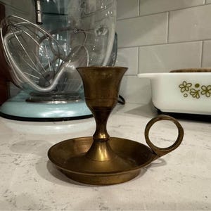 May include: A vintage brass candlestick holder. It has a round base, a cup-shaped top, and a small handle. The candlestick holder is made of brass and has a warm, golden colour. The design is simple and elegant, suitable for various decor styles.