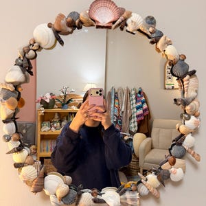 Puede incluir: Espejo redondo con un marco decorado con varias conchas marinas en tonos blancos, marrones y beige. El espejo refleja una habitación con una persona sosteniendo un teléfono, una estantería y un sillón beige.