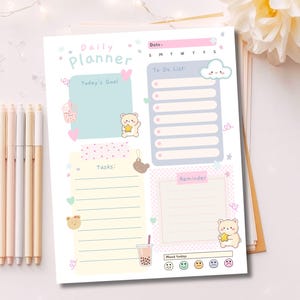 Könnte beinhalten: Ein Daily Planner-Blatt in Pastellfarben. Der Planer enthält Abschnitte für Aufgaben, Erinnerungen und Stimmungsverfolgung. Entzückende Bärenillustrationen und Herzen schmücken die Seite. Ein Satz Pastellstifte ist links zu sehen.