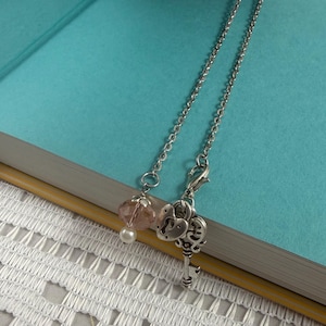 May include: A silver-toned metal chain necklace featuring a dangling charm set consisting of a faceted light pink crystal bead, a small white pearl bead, a heart-shaped padlock charm, and a detailed skeleton key charm attached with a lobster clasp.