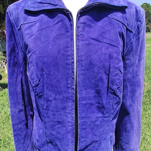 May include: A vibrant purple suede jacket featuring a full-length black front zipper, a structured collar, and long sleeves. The garment is constructed with visible seam detailing across the chest and shoulders, providing a tailored fit.