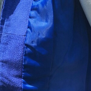 May include: A vibrant royal blue jacket featuring a textured suede-like outer material and a smooth, matching blue satin-finish lining. The garment includes a vertical zipper closure along the front edge, showcasing the contrast between the matte exterior.