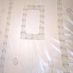 May include: A rectangular white tablecloth featuring a central rectangular panel of intricate crochet lace, a wide matching crochet lace border, and scattered circular crochet lace motifs across the smooth white fabric surface.