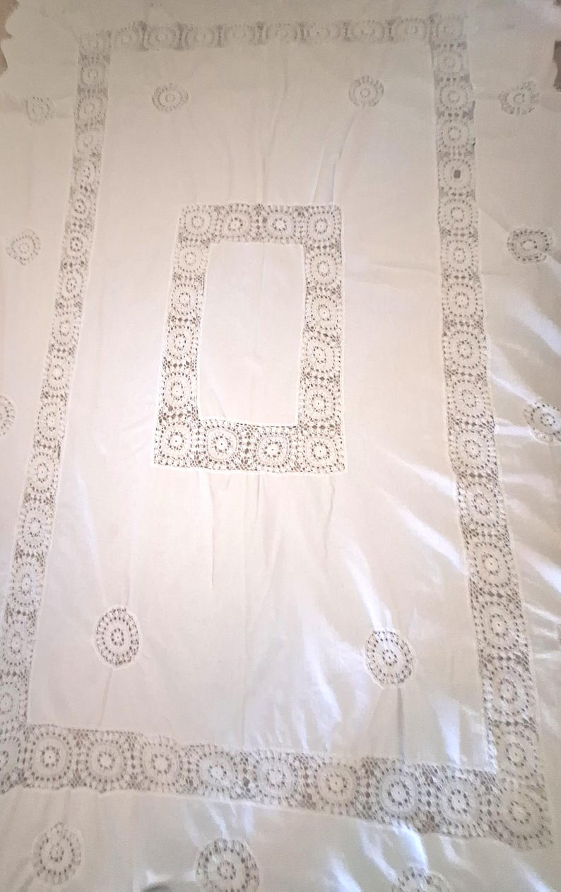 May include: A rectangular white tablecloth featuring a central rectangular panel of intricate crochet lace, a wide matching crochet lace border, and scattered circular crochet lace motifs across the smooth white fabric surface.