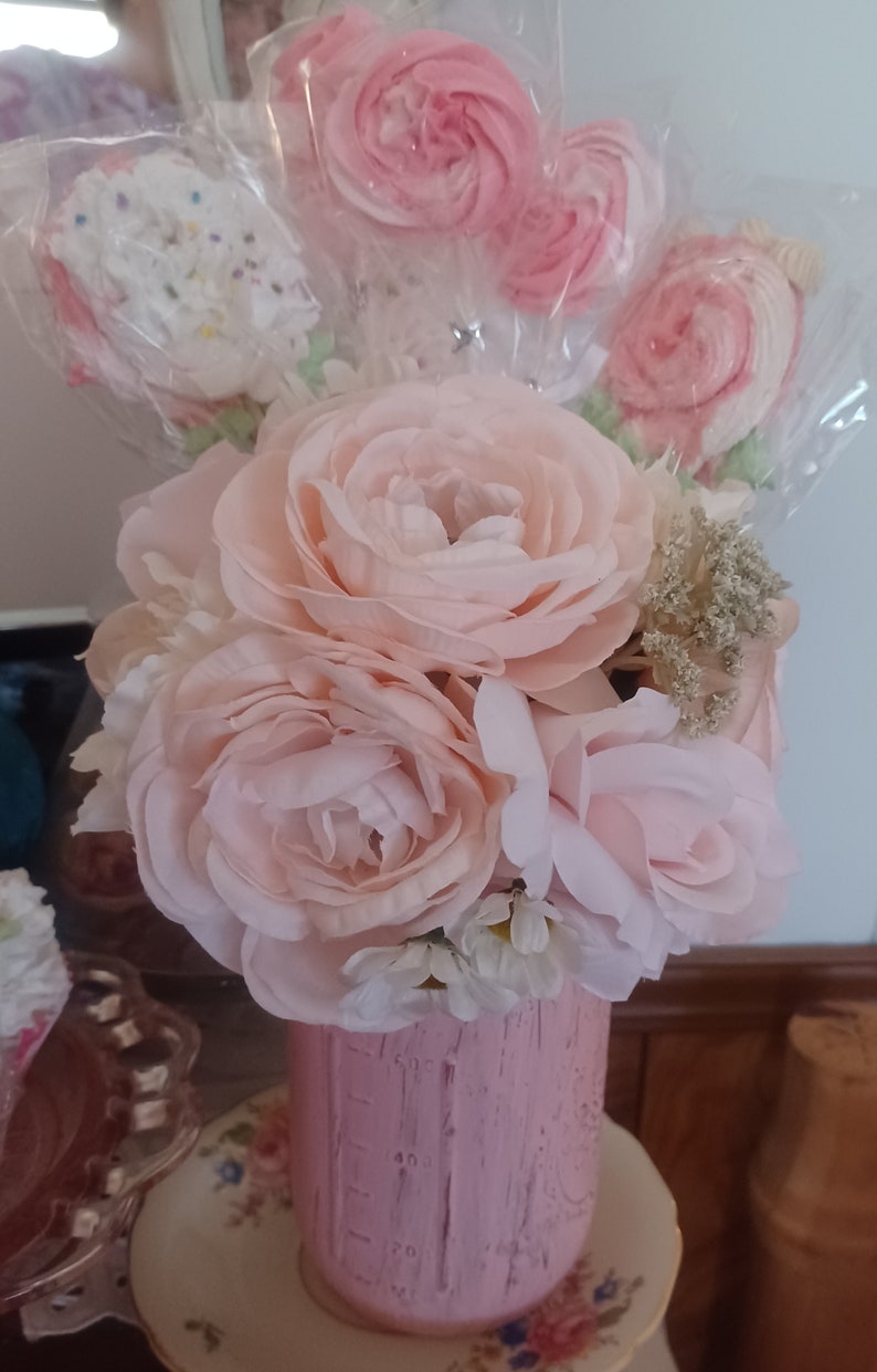 May include: A pink painted mason jar filled with light pink artificial roses and several meringue pops. The arrangement includes white and pink meringue pops on sticks, and a variety of faux flowers. The jar sits on a floral plate.