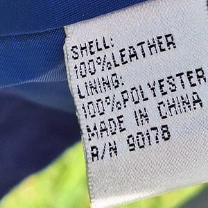 May include: A white rectangular garment care label with black text reading: SHELL: 100% LEATHER, LINING: 100% POLYESTER, MADE IN CHINA, R/N 90178. The label is stitched onto a blue fabric interior, with a portion of bright green fabric visible.