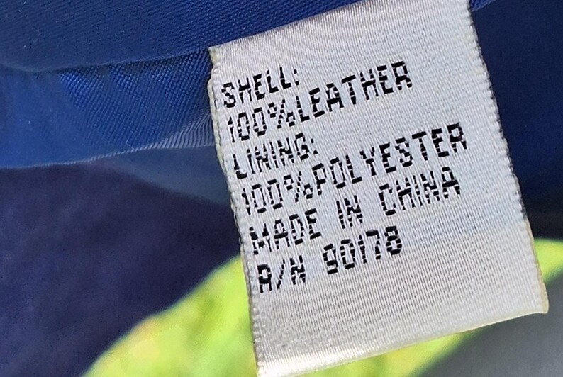 May include: A white rectangular garment care label with black text reading: SHELL: 100% LEATHER, LINING: 100% POLYESTER, MADE IN CHINA, R/N 90178. The label is stitched onto a blue fabric interior, with a portion of bright green fabric visible.