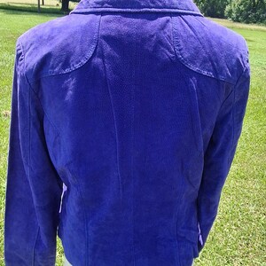May include: A vibrant purple suede jacket featuring a structured collar, long sleeves, and curved shoulder yoke detailing. The garment displays a textured finish across the back panel, with visible vertical seams extending from the yoke to the hem.