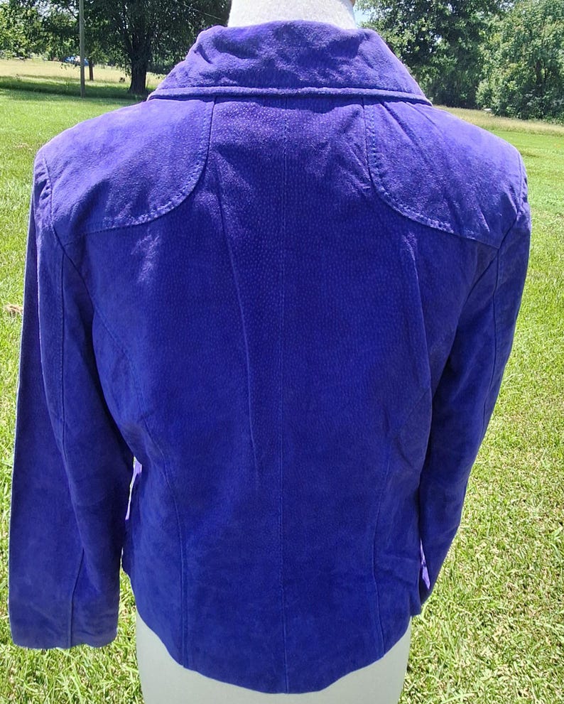 May include: A vibrant purple suede jacket featuring a structured collar, long sleeves, and curved shoulder yoke detailing. The garment displays a textured finish across the back panel, with visible vertical seams extending from the yoke to the hem.