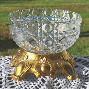 May include: A clear glass bowl featuring a faceted geometric pattern with a circular rim. The bowl is supported by a gold-colored metal base that has a wide, flared shape with embossed decorative details and three distinct feet.
