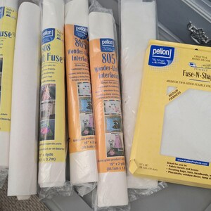 May include: A collection of Pellon brand sewing interfacing products, including several cylindrical rolls labeled 808 Craft-Fuse and 805 Wonder-Under, alongside a flat, rectangular yellow package of Fuse-N-Shape medium firm fusible interfacing.