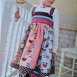May include: A colorful cupcake-themed dress with a white long-sleeved top. The dress features a brown bodice with pink and blue accents, and a skirt with various cupcake patterns. The text "the Cuppy Cake dress" is visible.