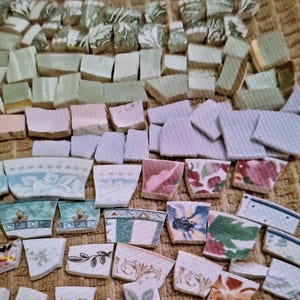 May include: A collection of assorted ceramic shards featuring various patterns and colors, including green floral prints, solid light green pieces, soft pink and lavender fragments, and detailed decorative motifs with blue, red, and gold accents.