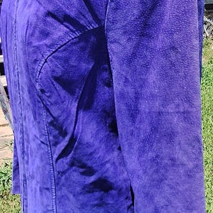 May include: A vibrant purple jacket featuring a textured finish, a high collar, and a full-length front zipper closure. The garment includes long sleeves and structured seam detailing along the front panels, providing a clean and fitted appearance.