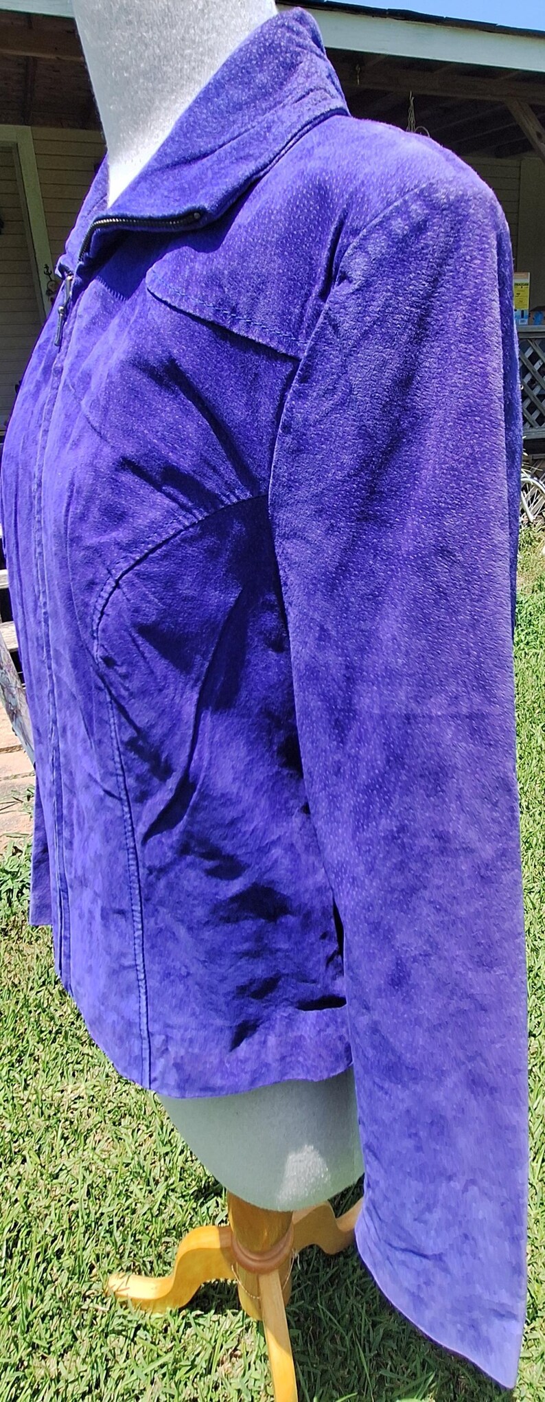 May include: A vibrant purple jacket featuring a textured finish, a high collar, and a full-length front zipper closure. The garment includes long sleeves and structured seam detailing along the front panels, providing a clean and fitted appearance.