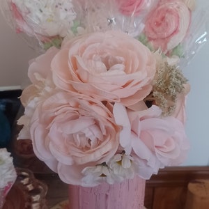 May include: A pink painted mason jar filled with light pink artificial roses and several meringue pops. The jar sits on a floral patterned plate. The meringue pops are wrapped in clear cellophane.