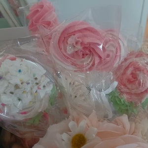 May include: Close-up of several pink and white meringue lollipops and cupcake-shaped treats, wrapped in clear cellophane. The lollipops are shaped like roses and swirls. The cupcake has white frosting with colorful sprinkles.